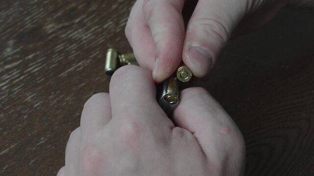 Close up of male hands loading brass bullets into handgun magazine. Metallic ammunition cartridges scattered on dark wooden background during firearm preparation.