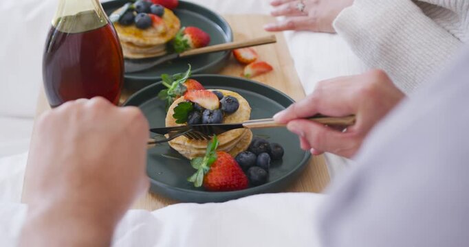 Foreground diner is placing fork and knife onto pancakes on bed tray, slicing bite to eat
