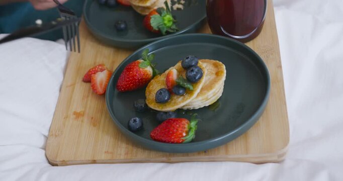 Hand entering from left is cutting pancakes with knife and fork, preparing bite on bed tray