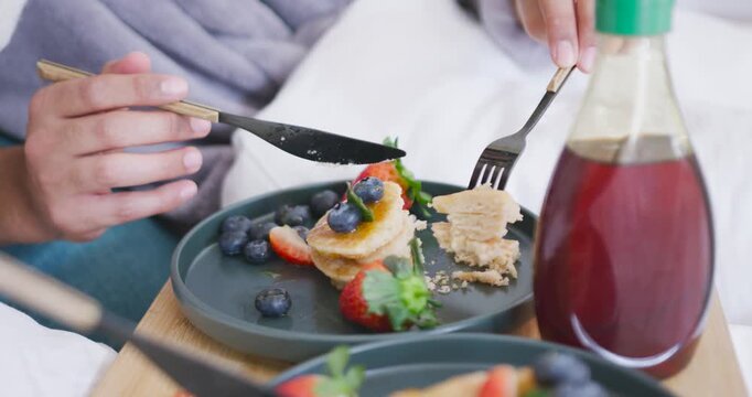Knife is cutting pancakes on plate on bed tray as fork is lifting bite for eating