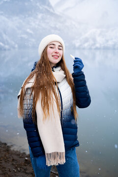 Young woman in warm winter clothing applying moisturizing lip balm while looking at the camera against a snowy mountain lake, representing winter lip care and beauty skincare.