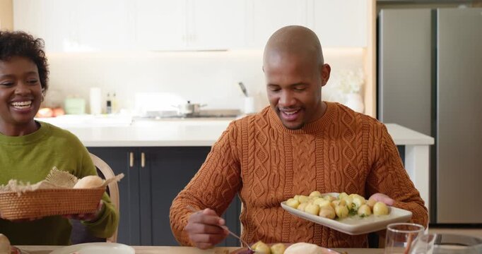 Family at kitchen table, adult man in rust sweater receiving platter and scooping potatoes for meal