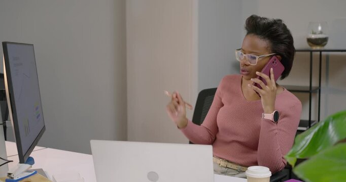 African American woman answering pink phone at desk, discussing charts and pointing pen at monitor