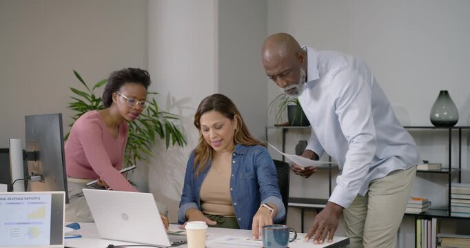 Diverse coworkers reviewing sales report on monitor at desk typing laptop pointing charts to decide