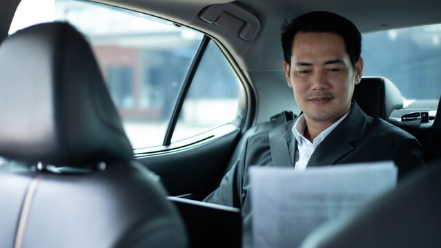 Asian businessman reading document while sitting in the back seat of a car, analyzing report on his commute, reflecting corporate executive lifestyle, mobility, and productivity during travel