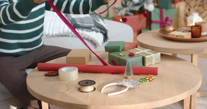 Person is reaching, pulling ribbon from spool to drape over box while wrapping gifts on table