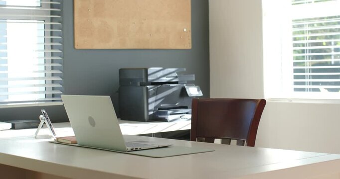 Sunlight is shifting through blinds as camera is panning to showcase tidy desk with laptop, printer