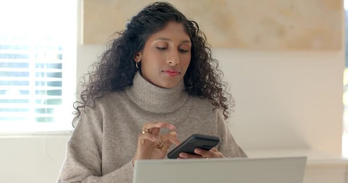 Female sitting at desk wearing turtleneck, speaking into smartphone while cross-referencing laptop
