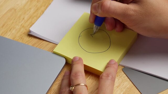 Person drawing a simple smiling face on a yellow sticky note using a blue ballpoint pen on a working desk