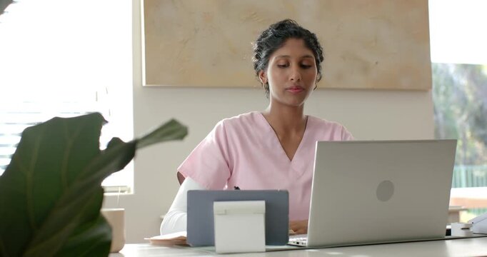 African American woman in scrubs at desk reading laptop checking tablet raising pen confirming info