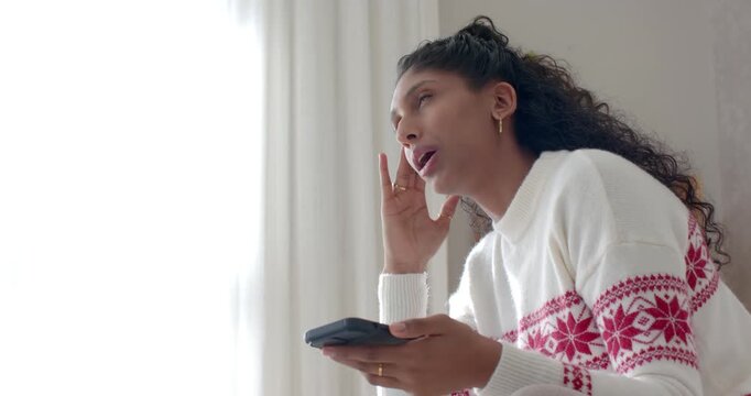 African American woman in white knit holding device responding to call while rubbing face by window