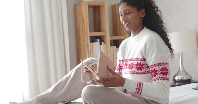 African American woman holding pink ribboned notebook flipping pages reading diary on bed by window