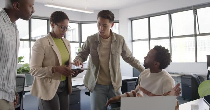 Diverse team in office leaning in pointing at laptop, woman in blazer tapping tablet solving issue