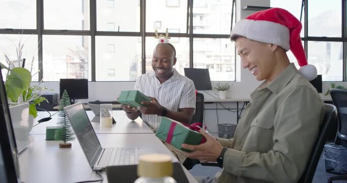During holiday Diverse coworkers exchanging green gifts at desk in office wearing Santa hat antlers