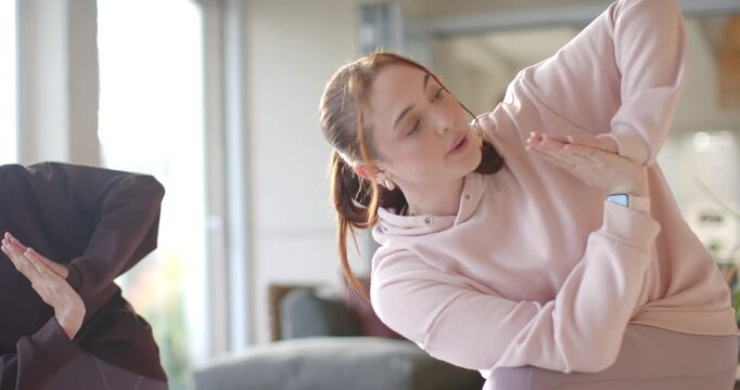 Female friends following cue, doing side-bend while breathing in living room with hoodie