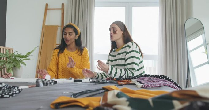 Diverse female friends showing jar and sorting clothing on bed, testing skincare patches chatting
