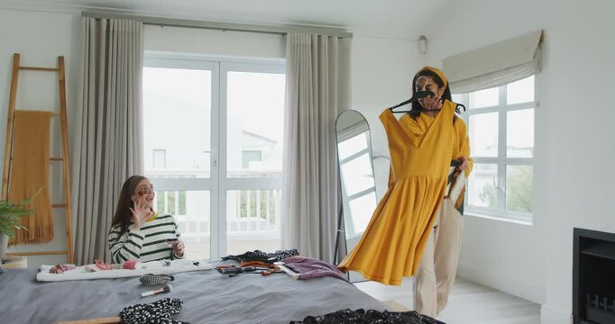 Diverse female friends sorting clothes on bed, holding yellow dress by mirror, preparing for outing