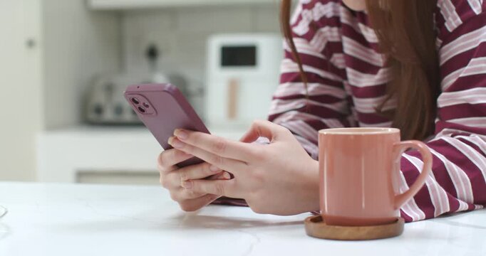 20s woman at kitchen counter holding mauve smartphone, tapping, checking messages, sipping pink mug