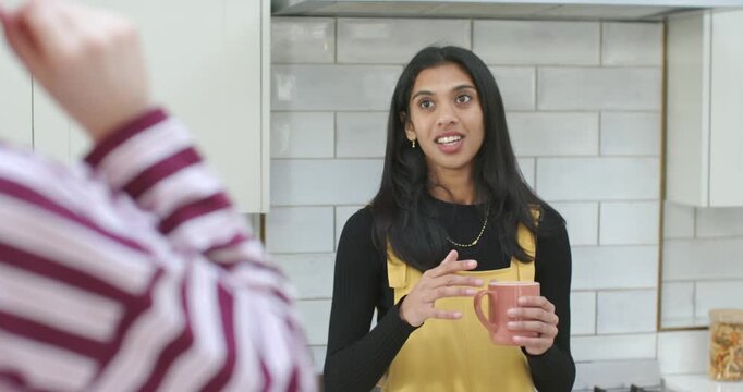 Indian woman at kitchen counter talking after housemate raising hand, wrapping pink mug and sipping