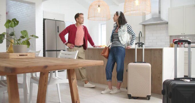 Couple standing and discussing travel plans after arriving in kitchen with beige suitcase smiling