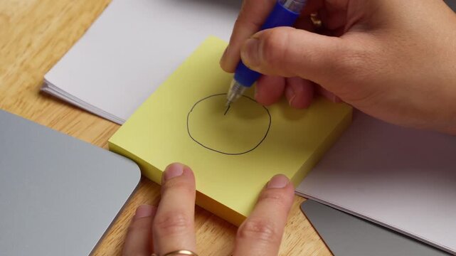Person drawing a simple sad face on a yellow sticky note using a blue ballpoint pen on a working desk