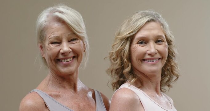 Two mature women wearing sleeveless tops after photographer cue, widening smiles in beige studio
