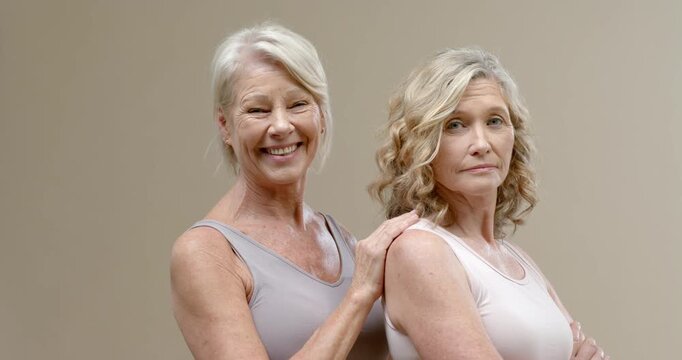 Senior female friends standing in tanks on tan backdrop left resting on shoulder showing support