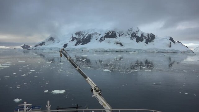 Panoramic view of Paradise Bay, Antarctica, from the stern of a cruise ship, with ship cranes in the foreground, calm waters, floating ice, snow-covered mountains and low clouds