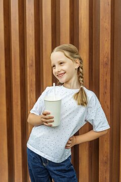Smiling Girl Holding Beverage Cup with Straw Against Wooden Wall