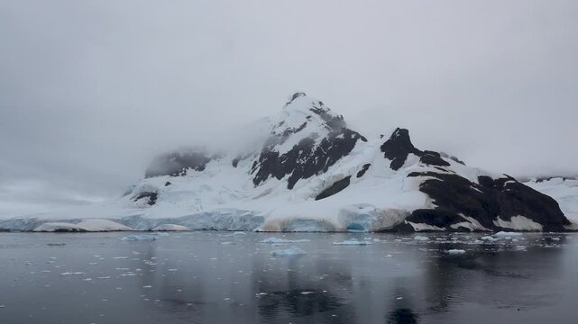 Scenic Antarctic coastline with snow-covered mountains and glaciers partly hidden by low clouds, reflected in calm waters with floating ice near Paradise Bay, penguins can be seen porpoising nearby