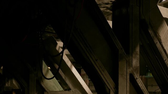 Workers are seen at a construction site in the evening. Lights and shadows create shapes within the structure while tools are scattered around the area. Activity persists despite the darkness.
