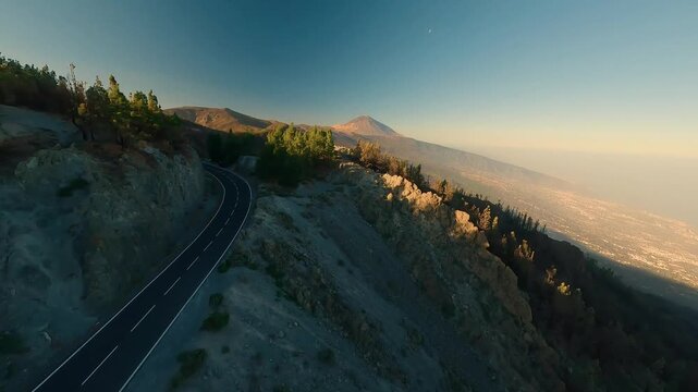 FPV aerial view of a winding road through a pine forest with Mount Teide in the background during golden hour in Santa Cruz de Tenerife, Canary Islands, Spain.