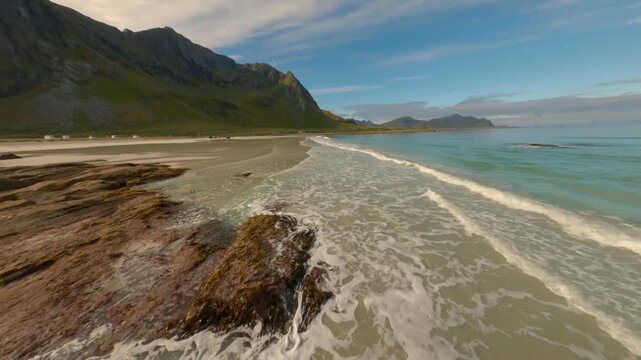 FPV aerial view of the sandy coastline and turquoise waters of Lofoten, with rocky outcrops and green mountains under a clear blue sky Nordland, Norway.