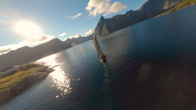 FPV aerial view of a traditional Viking ship sailing on the calm waters of a fjord in Lofoten, surrounded by dramatic mountains in Nordland, Norway.