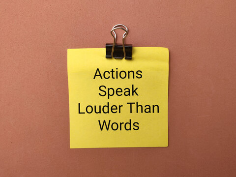 A binder clip secures a yellow note reading Actions Speak Louder Than Words on a brown surface