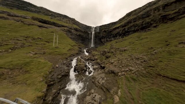 FPV aerial view of Fossa waterfall cascading down green grassy hills with a road bridge crossing the stream in the Faroe Islands.