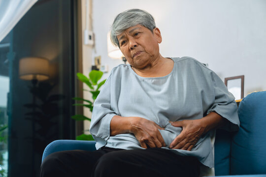 Elderly Asian woman hand holding stomach due to abdominal pain and discomfort sitting on a sofa at home
