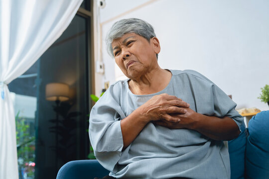 Elderly Asian woman holding her chest due to chest pain and discomfort and sitting on a sofa.