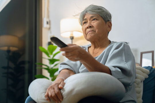 Elderly Asian woman relaxing on a sofa at home, holding a television remote while enjoying a favorite program.