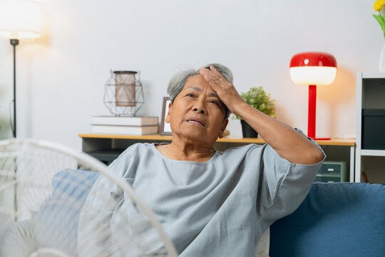Elderly Asian woman sitting on a sofa, feeling stressed from the heat while turning on a fan to cool down in an overheated living room.