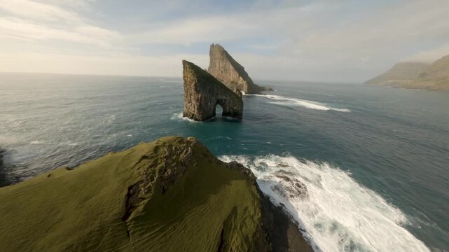 FPV aerial view of the iconic Drangarnir sea stacks with natural rock arch rising from the Atlantic Ocean along the rugged green coastline of the Faroe Islands.