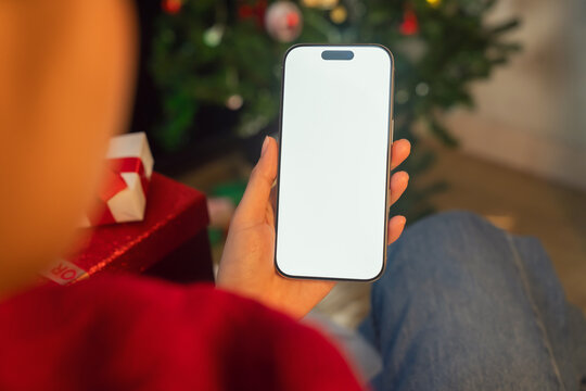 Hand holding a smartphone with a blank screen with gifts and a decorated tree, online shopping and Christmas celebration concept.