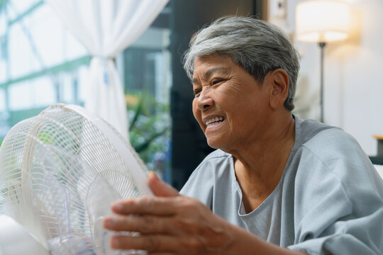 Cheerful elderly Asian woman sitting on a sofa and turning on a fan to cool down during extremely hot weather at home.