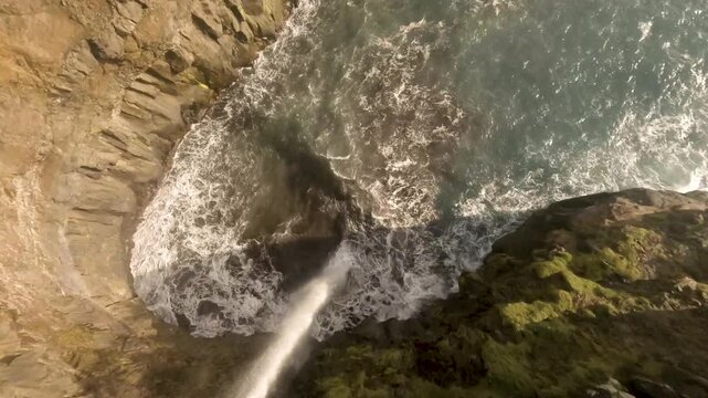 FPV aerial view of Mulafossur waterfall cascading down green cliffs into the ocean near Gasadalur village in the Faroe Islands.