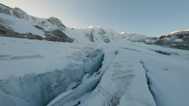 FPV aerial view of a deep crevasse in a snow-covered glacier with rugged mountain peaks under a clear sky in Meggen, Luzern, Switzerland.
