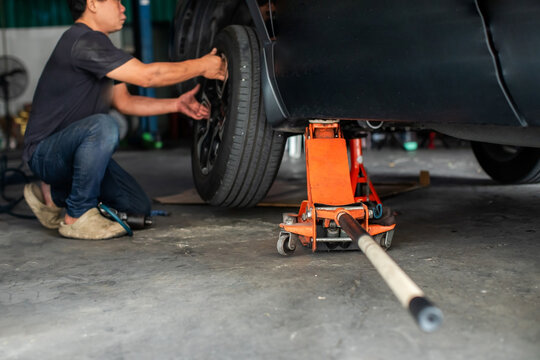 Selective focus to hydraulic floor jack with blurry professional mechanic installing a tire on a lifted car using an orange hydraulic floor jack and jack stand in a repair shop.