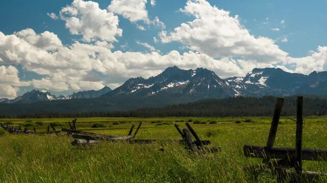 View of Sawtooth Mountains with a wooden fence in a green meadow under a blue sky in Stanley, Idaho, United States.