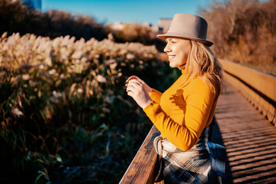 Happy woman in hat leaning on wooden pier &mdash; nature mindfulness and solo travel concept. Smiling female tourist enjoying sunny autumn afternoon near lake. Outdoor recreation, mental well-being.