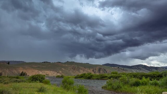 View of river flowing through a landscape with rolling hills under a dramatic, dark, stormy sky in Stanley, Idaho, United States.