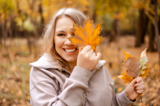 Happy woman covering eye with autumn leaf in forest &mdash; joyful seasonal recreation. Inner child healing and playful behavior pattern. Cheerful lady having fun outdoor connecting with peaceful nature.